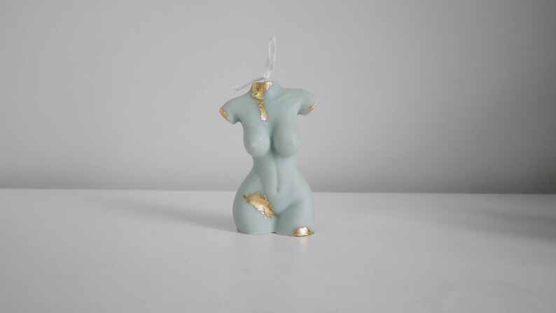Peut inclure: Une bougie en forme de torse bleu clair avec des accents de feuilles d'or. La bougie a une m&egrave;che en ficelle et est pr&eacute;sent&eacute;e sur une surface blanche. Un objet de d&eacute;coration unique.