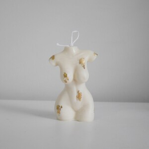 Peut inclure: Bougie en forme de torse de couleur ivoire avec des accents de feuilles d'or. La bougie a une m&egrave;che en haut et est plac&eacute;e sur une surface blanche. C'est un objet d&eacute;coratif, probablement pour la d&eacute;coration int&eacute;rieure ou les cadeaux.