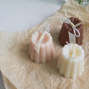 Peut inclure: Trois petites bougies rondes de différentes couleurs, marron, rose et blanc, avec un motif cannelé. Chaque bougie est ornée d'un ruban blanc noué en un nœud.