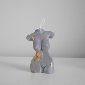 Peut inclure: Une bougie grise en forme de torse avec des accents de feuilles d'or. La bougie a une surface lisse et une m&egrave;che blanche. Il s'agit d'un article d&eacute;coratif, probablement pour la d&eacute;coration int&eacute;rieure ou comme cadeau.