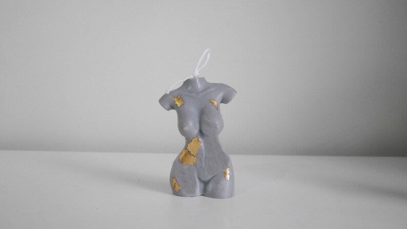 Peut inclure: Une bougie grise en forme de torse avec des accents de feuilles d'or. La bougie a une surface lisse et une m&egrave;che blanche. Il s'agit d'un article d&eacute;coratif, probablement pour la d&eacute;coration int&eacute;rieure ou comme cadeau.