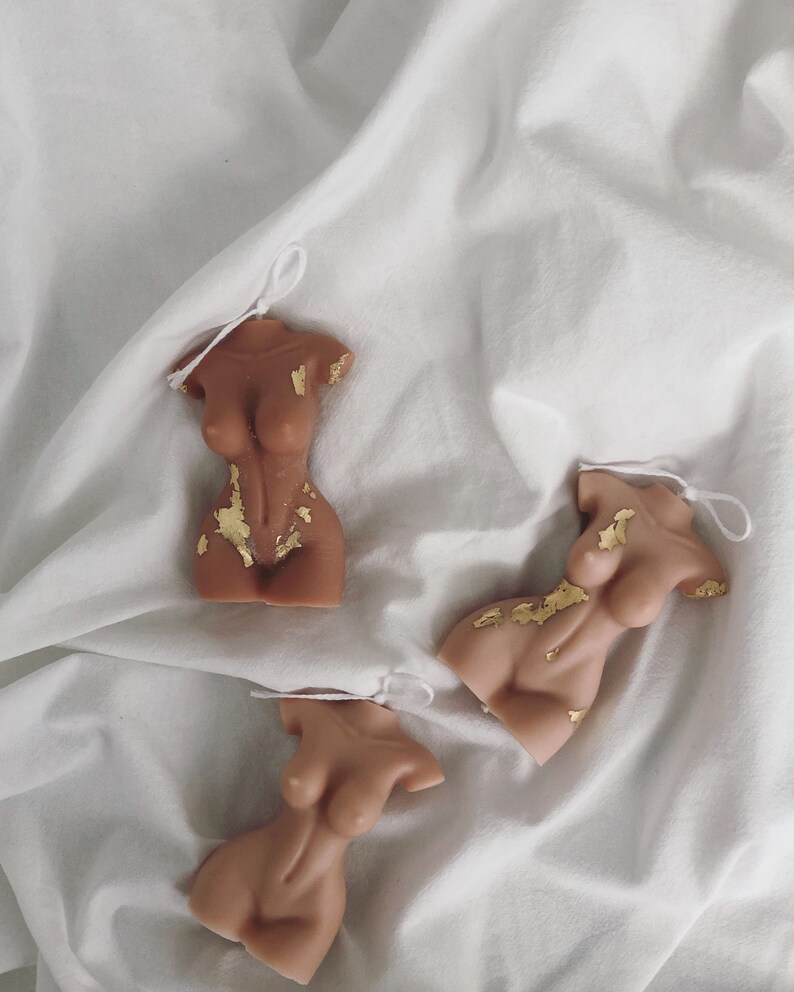 Peut inclure: Trois bougies en forme de torse de couleur nude, avec des accents de feuilles d'or et une ficelle blanche attach&eacute;e. Les bougies sont dispos&eacute;es sur un fond en tissu blanc. Les bougies ont la forme d'un corps f&eacute;minin.