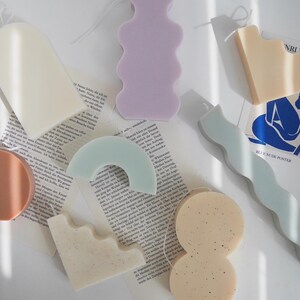 Peut inclure: Assortiment de bougies colorées de formes variées, notamment une forme ondulée, une forme arrondie et une forme rectangulaire. Les bougies sont posées sur une surface blanche avec un livre et une feuille de papier avec le texte "Henri Matisse" et "Blue Nude Poster".