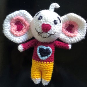 May include: A crocheted stuffed toy mouse with large white ears, a pink nose, and a red and yellow outfit. The mouse has a blue heart on its chest.