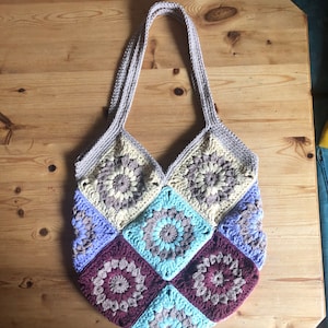 Könnte beinhalten: Eine gehäkelte Tragetasche mit Patchwork-Muster. Die Tasche besteht aus Quadraten in den Farben Lila, Blau, Braun und Beige. Die Tasche hat zwei lange Henkel.