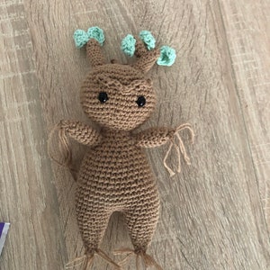 Pode incluir: Um brinquedo de amigurumi de cor marrom crochetado com forma humanoide e folhas verdes na cabeça. O brinquedo tem olhos pretos e um corpo marrom com pernas longas marrons.