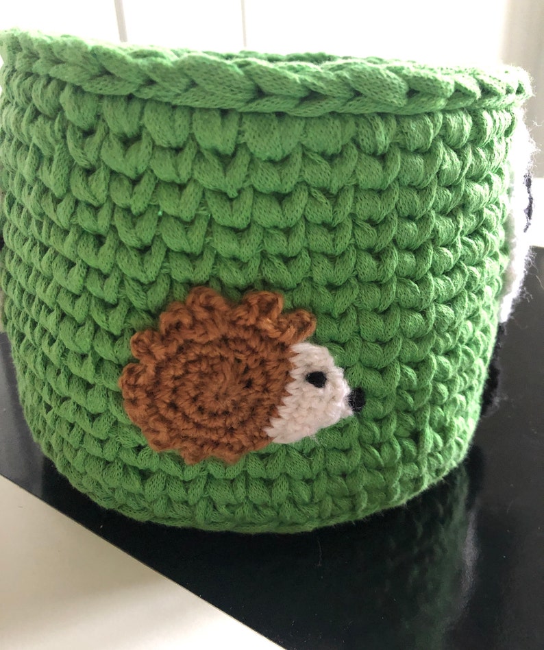 K&ouml;nnte beinhalten: Gr&uuml;ner H&auml;kel-Aufbewahrungskorb mit einem braunen und wei&szlig;en Igel-Applikationen.
