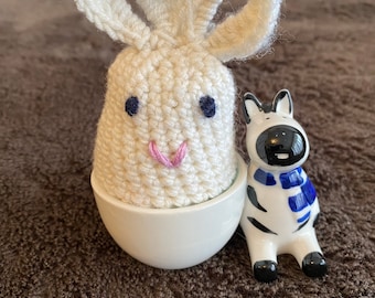 Könnte beinhalten: Ein weißer Häkelhase mit rosa Nase und schwarzen Augen sitzt in einem weißen Eierbecher. Eine schwarz-weiße Zebrafigur mit einem blauen Schal steht neben dem Eierbecher.