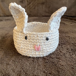 May include: White crocheted bunny basket with black eyes and a pink nose. The basket is on a brown fuzzy surface.
