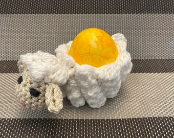 Könnte beinhalten: Ein gehäkelter weißer Lamm-Eierhalter mit einem gelben Osterei darin.