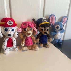 May include: Four handmade crochet character dolls. The dolls include a white dog with a red hat, a brown dog with pink goggles, a brown dog with a blue hat, and a blue alien with large ears. The dolls are sitting on a white surface.