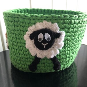 K&ouml;nnte beinhalten: Gr&uuml;ner H&auml;kelkorb mit einer schwarzen und wei&szlig;en Schaf-Applikation. Das Schaf hat Glitz Augen und eine schwarze Nase.