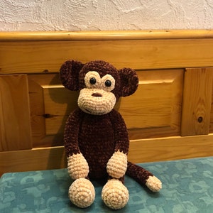 Op de afbeelding: Een bruin gehaakt aapje met beige handen en voeten. De aap heeft zwarte ogen en een bruine neus. Het speelgoed zit op een blauw gepatroneerd oppervlak.