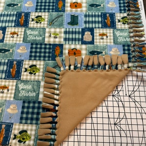 May include: A tan and blue fleece blanket with a "Gone Fishin'" theme. The blanket has a patchwork design with images of fish, fishing rods, boots, and fishing vests. The blanket has a fringe border.