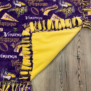 May include: Purple and gold fleece blanket with a Minnesota Vikings theme. The blanket has a fringe border and features the team logo and the words "Vikings" and "Minnesota".
