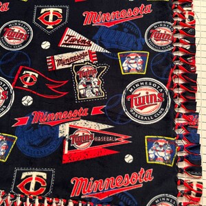 May include: A blue and red fleece blanket with a Minnesota Twins baseball theme. The blanket features the team's logo, the words "Minnesota Twins", and baseballs. The blanket has a knotted fringe border.