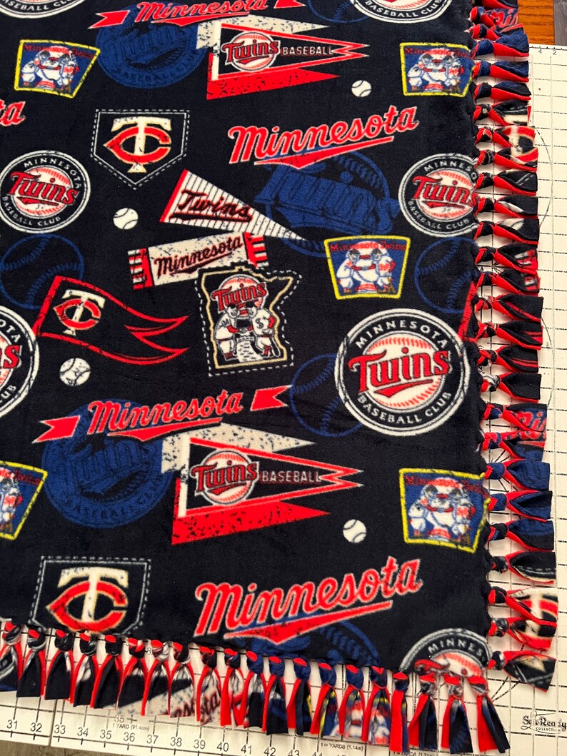 May include: A blue and red fleece blanket with a Minnesota Twins baseball theme. The blanket features the team's logo, the words "Minnesota Twins", and baseballs. The blanket has a knotted fringe border.