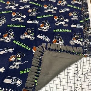 K&ouml;nnte beinhalten: Marineblaue Fleece-Decke mit einem sich wiederholenden Muster eines Zeichentrickcharakters in Football-Uniform, dem Seattle Seahawks-Logo und den Worten "Seattle Seahawks" in Gr&uuml;n und Wei&szlig;. Die Decke hat eine graue R&uuml;ckseite und einen geknoteten Fransenrand.