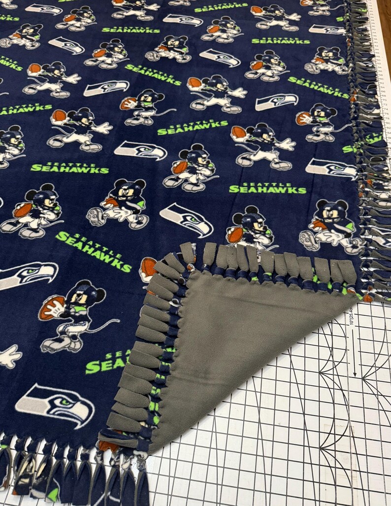 K&ouml;nnte beinhalten: Marineblaue Fleece-Decke mit einem sich wiederholenden Muster eines Zeichentrickcharakters in Football-Uniform, dem Seattle Seahawks-Logo und den Worten "Seattle Seahawks" in Gr&uuml;n und Wei&szlig;. Die Decke hat eine graue R&uuml;ckseite und einen geknoteten Fransenrand.