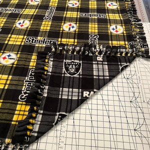 May include: A black and yellow fleece blanket with the word "Steelers" repeated throughout. The blanket has a fringe border and is folded over a black and white plaid fabric with the word "RAIDERS" and a Raiders logo.