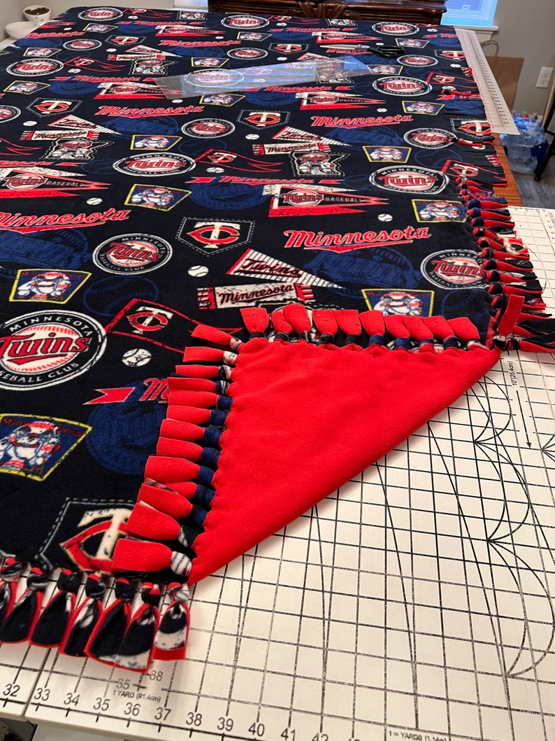 May include: A red and blue fleece blanket with a Minnesota Twins theme. The blanket has a fringe border and features a variety of logos and designs, including the team's logo, the words "Minnesota Twins", and baseballs. The blanket is folded in half and is lying on a white surface with a grid pattern.