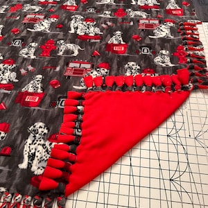 May include: A red and grey fleece tie blanket with a pattern of dalmatian dogs and fire trucks. The blanket has a fringe border and is folded in half.