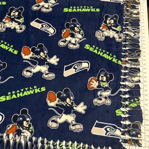 K&ouml;nnte beinhalten: Marineblaue Fleece-Decke mit einem sich wiederholenden Muster von Mickey Mouse in einem Football-Trikot der Seattle Seahawks, der einen Football h&auml;lt. Die Decke zeigt auch das Logo des Teams und die Worte "SEATTLE SEAHAWKS". Die R&auml;nder sind mit geknoteten Fransen versehen.