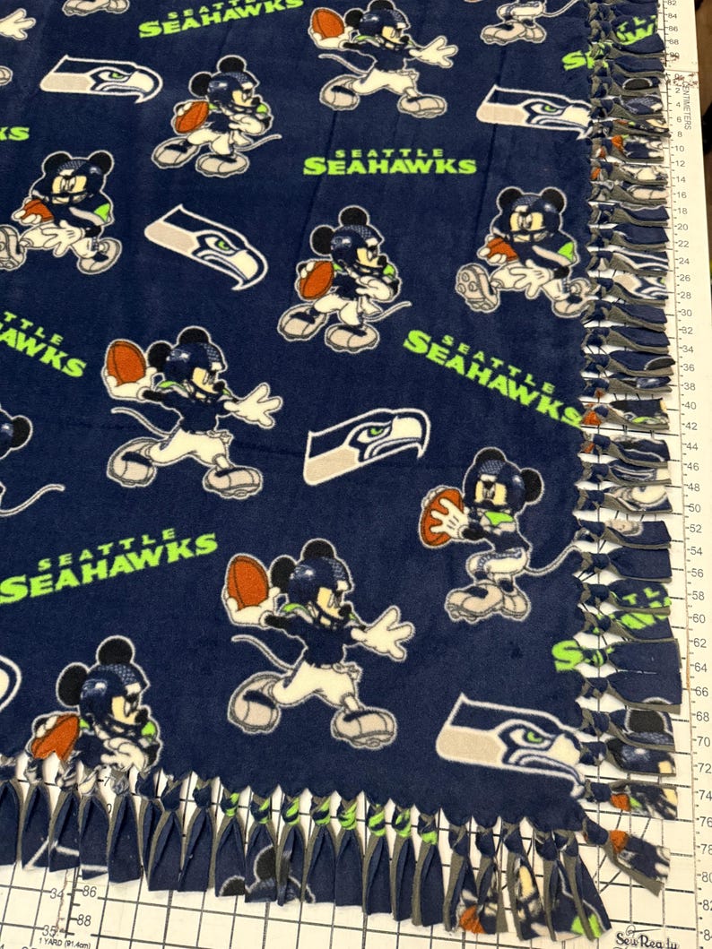 K&ouml;nnte beinhalten: Marineblaue Fleece-Decke mit einem sich wiederholenden Muster von Mickey Mouse in einem Football-Trikot der Seattle Seahawks, der einen Football h&auml;lt. Die Decke zeigt auch das Logo des Teams und die Worte "SEATTLE SEAHAWKS". Die R&auml;nder sind mit geknoteten Fransen versehen.