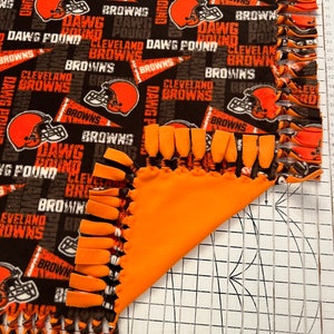May include: A brown and orange fleece blanket with a Cleveland Browns theme. The blanket has a fringe border and features the team's logo and the words "Dawg Pound" and "Cleveland Browns".