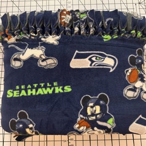 K&ouml;nnte beinhalten: Eine marineblaue Fleece-Decke mit dem Logo der Seattle Seahawks und Cartoon-Charakter-Grafiken. Die Decke hat einen geknoteten Fransenrand in verschiedenen Farben. Der Text "SEATTLE SEAHAWKS" ist gr&uuml;n gedruckt.