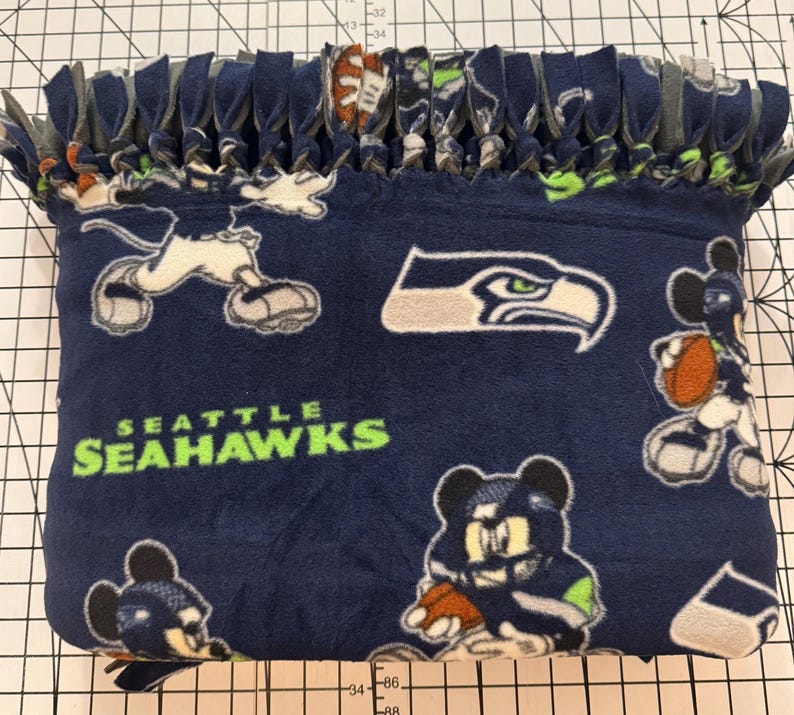 K&ouml;nnte beinhalten: Eine marineblaue Fleece-Decke mit dem Logo der Seattle Seahawks und Cartoon-Charakter-Grafiken. Die Decke hat einen geknoteten Fransenrand in verschiedenen Farben. Der Text "SEATTLE SEAHAWKS" ist gr&uuml;n gedruckt.