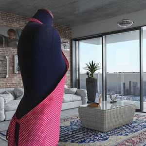 A pink and white polka dot apron. The apron is tied at the waist and has a long strap that goes over the shoulder. The apron is displayed on a mannequin in a modern living room setting.