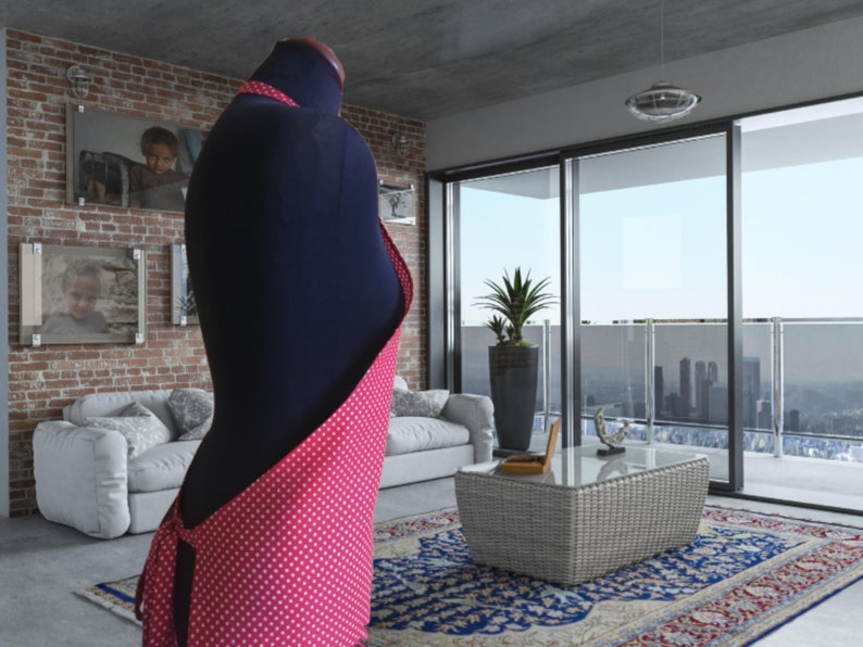 A pink and white polka dot apron. The apron is tied at the waist and has a long strap that goes over the shoulder. The apron is displayed on a mannequin in a modern living room setting.