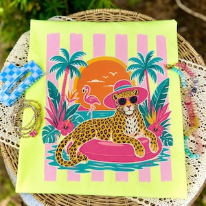 Peut inclure: Un t-shirt jaune néon avec un léopard portant des lunettes de soleil et un chapeau rose, se prélassant sur un anneau gonflable rose. Le graphisme comprend un coucher de soleil, des palmiers et un flamant rose. Accessoires : bracelets et pince à cheveux à carreaux bleus.
