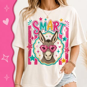 Peut inclure: T-shirt crème avec un âne de dessin animé portant des lunettes de soleil en forme de cœur. Le mot "SMART" est au-dessus de l'âne en lettres colorées. Des étoiles et des rayures roses et turquoise décorent le motif.