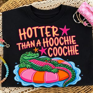 Peut inclure: T-shirt noir avec l'inscription "HOTTER THAN A HOOCHIE COOCHIE" en rose et orange. Le motif représente un alligator de dessin animé sur un bouée gonflable. Un accessoire pour cheveux rose et un collier de perles sont également visibles.