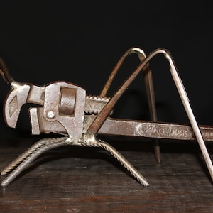 Op de afbeelding: Een roestig metalen sculptuur van een sprinkhaan gemaakt van een pijpsleutel en andere metalen onderdelen. Het sculptuur staat op een houten oppervlak.