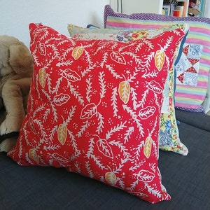 May include: Red cushion with a white and yellow floral pattern. The cushion is on a grey sofa with other cushions behind it.