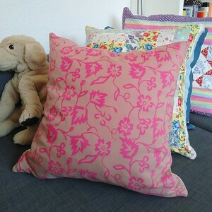 May include: A pink floral patterned cushion with a light brown background. The cushion is on a sofa with other cushions in the background.