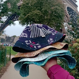 Puede incluir: Una pila de cuatro sombreros de cubo de tela en varios patrones y colores. El sombrero superior es negro con rayas blancas y un patrón ikat rosa y blanco. El segundo sombrero es un estampado floral dorado y marrón. El tercer sombrero es un estampado floral azul claro y blanco. El sombrero inferior es un estampado geométrico azul verdoso y blanco. Los sombreros se sostienen frente a la Torre Eiffel.