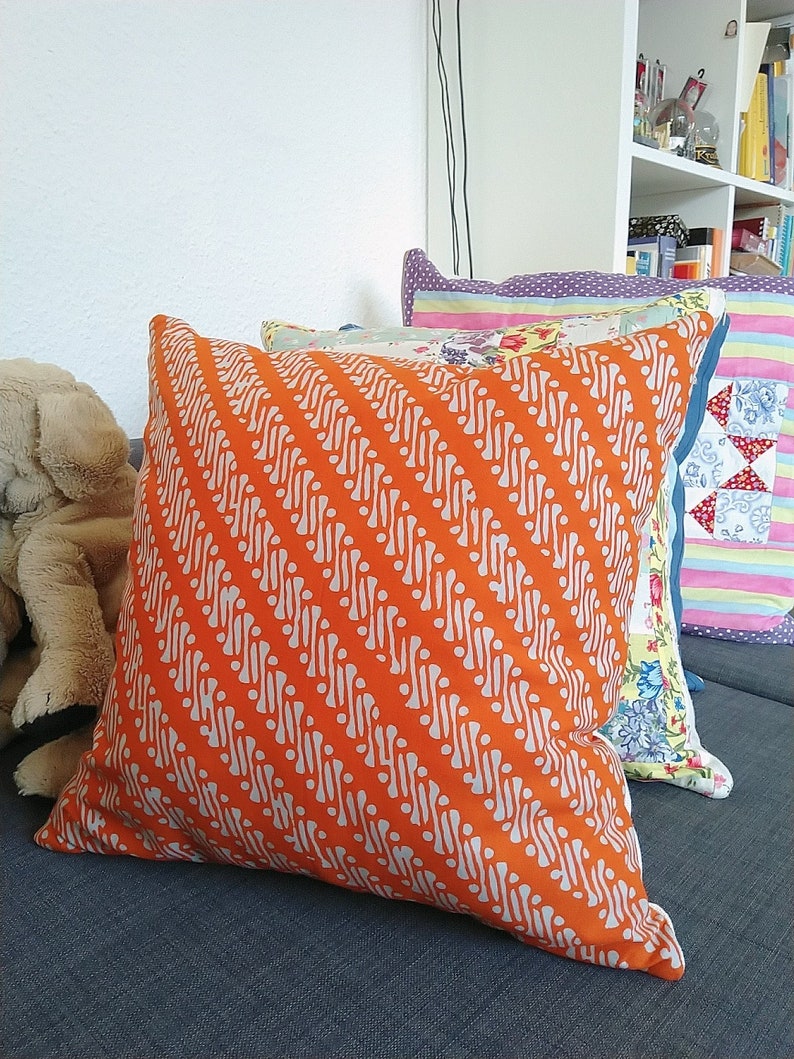 May include: A set of three decorative cushions with different patterns. The largest cushion is orange with a white geometric pattern. The middle cushion is white with a floral pattern. The smallest cushion is purple with white polka dots.