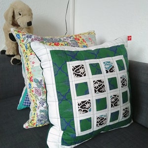 May include: Three decorative pillows on a gray couch. The pillow in the foreground is green with a white border and a geometric pattern. The other two pillows are made of patchwork fabric with floral and geometric patterns.