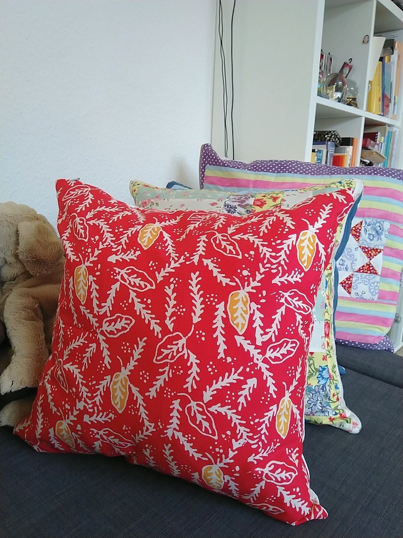 Pu&ograve; includere: Cuscino rosso con un motivo floreale bianco e giallo. Il cuscino &egrave; su un divano grigio con altri cuscini dietro.