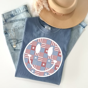 May include: A blue t-shirt with a white smiley face design. The smiley face is made up of red, white, and blue patchwork squares. The shirt has the text "COMFY COLORS" on the tag.