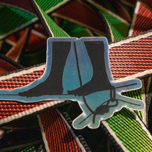 May include: A silver sticker with a black silhouette of two legs wearing socks and standing on a rope. The sticker is on top of a red, white, and green striped fabric.