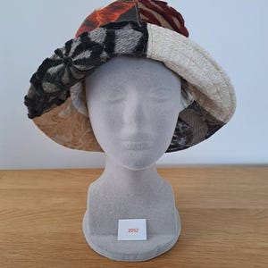 May include: A patchwork bucket hat in various textured fabrics, including brown, black, grey, and cream. The hat is displayed on a mannequin head. The tag on the base of the mannequin reads "2052".
