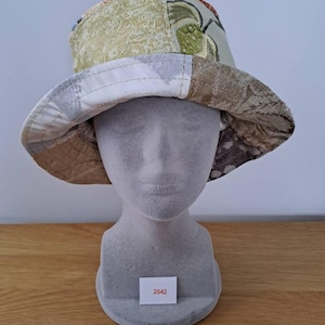 May include: A patchwork bucket hat with a wide brim. The hat features fabrics in green, beige, and brown. The brim is lined with white fabric with a grey pattern. The hat is displayed on a mannequin head.