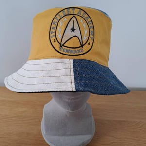May include: A yellow, white, and blue bucket hat featuring the Starfleet Academy Command logo. The hat is made from denim and cotton. The brim is white and blue, with a stitched design.