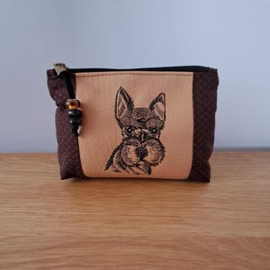 May include: A small, brown zippered pouch featuring an embroidered Schnauzer dog. The pouch has a tan central panel and brown polka-dot side panels. A beaded charm is attached to the zipper pull.