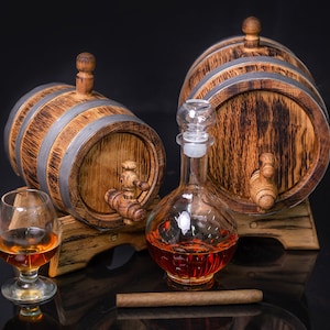 May include: Two wooden barrels with metal bands and spigots, a glass decanter filled with amber liquid, a glass of amber liquid, and a brown cigar on a black surface.